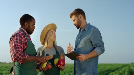 Young-Pretty-Woman-And-Two-Multiethnic-Men-Farmers-Standing-In-The-Green-Field-With-Harvested-Vegetables-In-Hands-And-One-Man-With-A-Tablet-Device