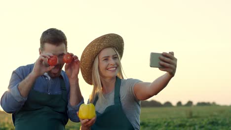 Fröhliche-Junge,-Attraktive-Mann--Und-Frauenbauern,-Die-In-Die-Smartphone-Kamera-Lächeln-Und-Selfie-Fotos-Auf-Dem-Feld-Mit-Gemüse-In-Den-Händen-Machen-Und-Spaß-Mit-Ihnen-Haben