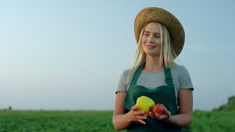 Portrait-Of-The-Pretty-Young-Blond-Woman-Farmer-In-A-Hat-And-Apron-Standing-In-The-Green-Field-And-Posing-To-The-Camera-With-Harvested-Vegetables-In-Hands-Then-Smelling-Them