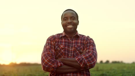 Portrait-Of-The-Handsome-Young-Man-Farmer-Standing-In-The-Field-On-The-Sunset,-Smiling-To-The-Camera-And-Crossing-His-Hands-In-Front-Of-Him