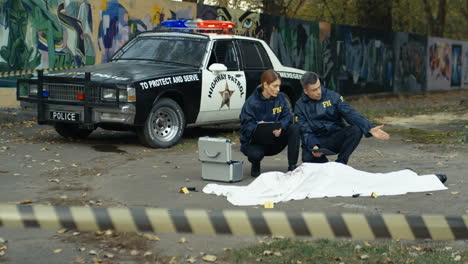 Couple-Of-Police-Officers-Sitting-And-Talking-Over-The-Dead-Body-Lying-On-Street-Under-White-Sheet