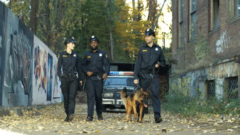 Drei-Multiethnische-Polizisten,-Die-Mit-Einem-Schäferhund-Die-Alte-Gasse-Entlanggehen-Und-Etwas-Suchen