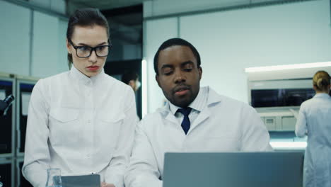 Male-Worker-Of-The-Laboratory-Sitting-At-The-Computer-And-Checking-Something-While-His-Young-Female-Assistant-Standing-With-A-Tablet-Device-Next-To-Him