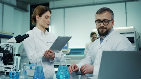 Medical-Laboratory-Male-And-Female-Scientists-Talking-About-Their-Researching-And-Discovery-While-Man-Sitting-At-The-Laptop-Computer-And-Woman-Standing-With-A-Folder