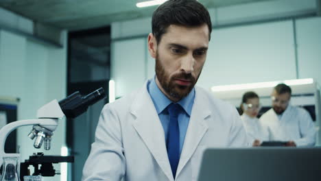 Close-Up-Of-The-Attractive-Man-Working-In-The-Laboratory-And-Looking-In-The-Microscope,-Then-Typing-On-The-Laptop-Computer