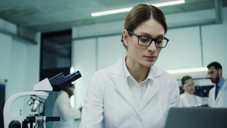 Close-Up-Of-The-Beautiful-Young-Woman-In-Glasses-Working-In-The-Laboratory-And-Looking-In-The-Microscope,-Then-Typing-On-The-Laptop-Computer