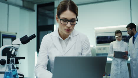 Porträt-Der-Gut-Aussehenden-Frau-Mit-Brille-Und-Weißem-Gewand,-Die-Am-Laptop-Computer-Untersucht-Und-Analysiert,-Während-Sie-Im-Labor-In-Das-Mikroskop-Schaut