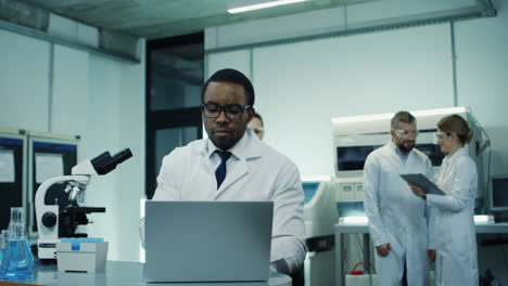 Young-Man-Scientist-In-The-White-Robe-And-Glasses-Working-At-The-Laptop-And-Microscope-Over-Research-While-His-Female-Co-Worker-Coming-With-A-Tablet-And-They-Discussing-Some-Results