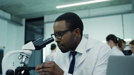 Close-Up-Of-The-Young-Attractive-Man,-Medical-Researcher,-Looking-In-The-Microscope-While-Doing-Some-Analysis-And-Typing-Them-In-The-Laptop-Computer-At-The-Laboratory