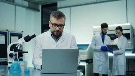 Nahaufnahme-Des-Mannes-Mit-Brille,-Medizinischer-Forscher,-Der-In-Das-Mikroskop-Schaut,-Während-Er-Einige-Analysen-Durchführt-Und-Sie-In-Den-Laptop-computer-Im-Labor-Eingibt