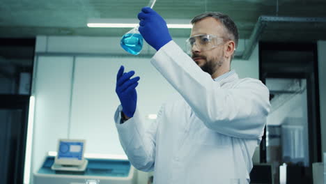 Camera-Zooming-In-On-The-Male-Laboratory-Scientist-In-The-Glasses-And-White-Robe-Making-An-Experiment-Or-Analysis-With-A-Blue-Liquid-In-The-Test-Tube