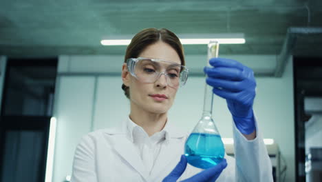 Camera-Zooming-Out-From-The-Woman-In-Glasses-And-White-Robe-Doing-An-Analysis-Of-The-Blue-Liquid-In-The-Test-Tube-During-Medical-Or-Pharmaceutical-Research-And-In-The-Laboratory