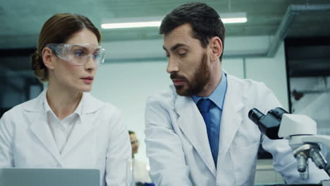 Portrait-Of-The-Two-Laboratory-Scientists,-Man-And-Woman,-Talking-While-He-Working-At-The-Microscope-And-She-At-The-Laptop-Computer