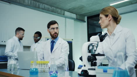 Woman-Scientist-In-The-White-Robe-Looking-In-The-Microscope-While-Doing-Some-Investigation-And-Her-Male-Colleague-Testing-Some-Liquid-In-Tube-At-The-Laptop-Computer