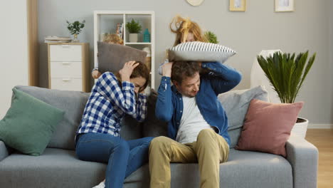 Young-Parents-Sitting-On-The-Sofa-In-The-Living-Room-And-Their-Funny-Cheerful-Kids-Beating-Them-With-Pillows