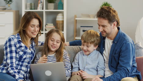 Joyful-Young-Parents-With-Little-Daughter-And-Son-Sitting-On-The-Sofa-In-The-Living-Room-And-Talking-While-Having-Videochat-On-The-Laptop-Computer