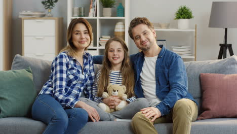 Portrait-Shot-Of-The-Young-Happy-Parents-Sitting-With-Their-Little-Cute-Daughter-On-The-Couch-In-The-Living-Room,-Looking-At-Eacj-Other-And-Smiling-To-The-Camera