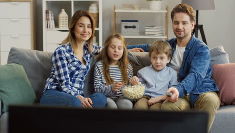 Junge-Eltern-Mit-Ihren-Kindern-Sitzen-Auf-Der-Couch-Im-Wohnzimmer,-Essen-Popcorn-Und-Fernsehen