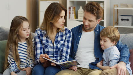 Cheerful-Family-Of-Parents-And-Little-Son-And-Daughter-Sitting-All-Together-On-The-Couch-And-Flipping-The-Pages-Of-The-English-Book