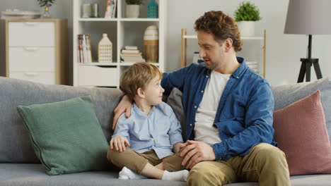 Young-Handsome-Father-Sitting-On-The-Sofa-In-The-Living-Room-And-Talking-With-His-Small-Cute-Son-Who-Sitting-Next-To-Him