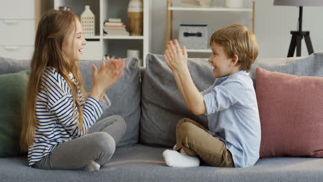 Two-Small-Cute-Kids,-Sister-And-Brother,-Sitting-On-The-Couch-With-Pillows-In-The-Cozy-Room-And-Playing-A-Game-With-Their-Hands-By-Clapping-Them