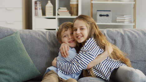 Portrait-Shot-Of-The-Cute-Small-Sister-And-Brother-Sitting-On-The-Couch-In-The-Living-Room,-Hugging-Each-Other-And-Posing-In-Front-Of-The-Camera