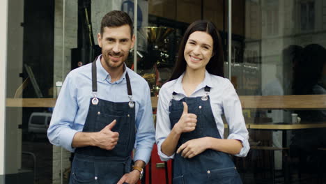 Young-Cheerful-Waiter-And-Waitress-Turning-To-The-Camera,-Giving-Their-Thumbs-Up-And-Smiling-While-Standing-At-The-Entrance-To-The-Restaurant