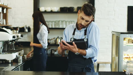 Attractive-Waiter-Speaking-On-The-Telephone-And-Using-Tablet-Computer-At-The-Bar,-Brunette-Waitress-Making-Coffee-Behind
