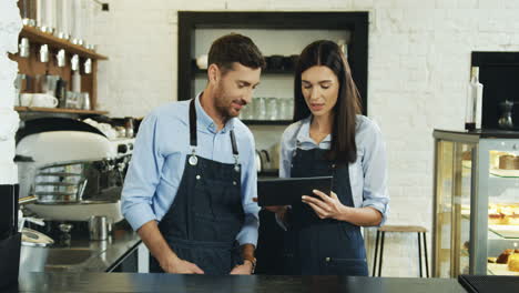Attractive-Waitress-And-Waiter-Smiling-And-Watching-Something-On-The-Tablet-Computer-While-Standing-At-The-Bar-In-The-Cafe