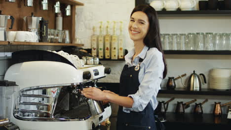 Porträtaufnahme-Der-Gut-Aussehenden-Brünetten-Kellnerin,-Die-Auf-Der-Kaffeemaschine-Kaffee-Mit-Milch-Kocht-Und-In-Die-Kamera-Lächelt