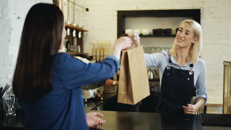 Woman-Buying-Something-At-The-Bar-In-The-Cafe-And-Pretty-Blonde-Waitress-Giving-Her-A-Cartoon-Bag,-Then-She-Going-Away