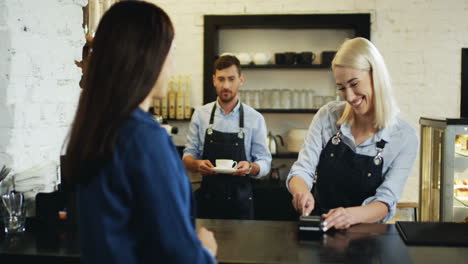 Rear-Of-The-Brunette-Woman-Paying-With-A-Credit-Card-To-The-Blonde-Pretty-Waitress-At-The-Bar,-While-Waiter-Bringing-Her-Coffee