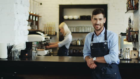 Portrait-Shot-Of-The-Handsome-Smiled-Waiter-Standing-And-Leaning-On-The-Bar-While-Blond-Attractive-Waitress-Preparing-Coffe-Behind