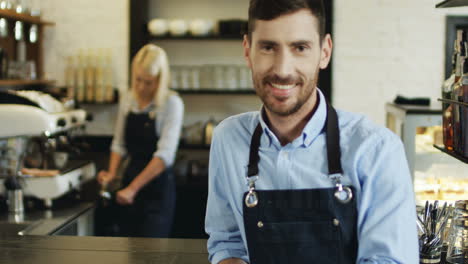 Portrait-Of-The-Good-Looking-Smiled-Barmen-Standing-At-The-Bar-While-Blond-Young-Waitress-Making-Coffe-On-The-Background