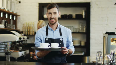 Portrait-Shot-Of-The-Handsome-Barmen-Holding-Cups-Of-Coffee-And-Going-To-The-Bar-While-His-Female-Co-Worker-Preparing-More-Behind-At-The-Coffee-Machine