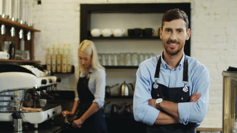 Portrait-Of-The-Young-Handsome-Waiter-Standing-In-Front-Of-The-Camera-And-Watching-Behind-At-The-Young-Blond-Waitress-Making-Coffee-On-The-Special-Machine,-Then-Smiling-To-The-Camera