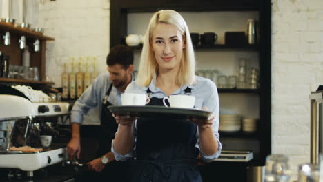 Portrait-Shot-Of-The-Beautiful-Waitress-Holding-Cups-Of-Coffee-And-Going-To-The-Bar-While-Her-Male-Co-Worker-Preparing-More-Behind-At-The-Coffee-Machine