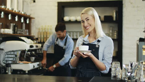 Portrait-Shot-Of-The-Young-Charming-Waitress-Standing-In-Front-Of-The-Camera-With-Cup-Of-Coffee-In-Hands-And-Watching-Behind-At-The-Waiter-Near-Coffee-Machine,-Then-Smiling-To-The-Camera