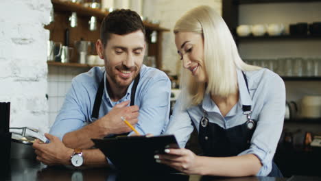Young-Good-Looking-Waiter-And-Waitress-Standing-At-The-Bar-And-Making-Menu-Set,-Woman-Writing-With-Pencil