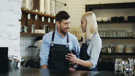 Cheerful-Good-Looking-Waiter-And-Waitress-Standing-At-The-Bar,-Talking-And-Watching-Some-Video-On-The-Tablet-Computer