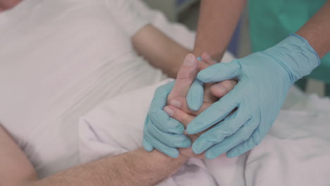 Detail-Of-A-Nurse's-Hands-With-Gloves-Grasping-And-Comforting-The-Hands-Of-A-Sick-Patient-On-Hospital-Bed