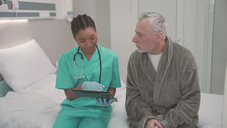 A-Beautiful-Young-American-Female-Doctor-Sitting-On-The-Edge-Of-A-Hospital-Bed-With-An-Old-Patient