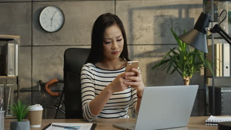 Beautiful-Young-Woman-Sitting-At-The-Table-With-Laptop-Computer-In-The-Office-And-Tapping-Nd-Texting-On-The-Smartphone-With-A-Smile