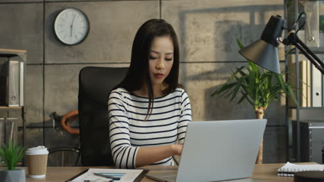 Young-Female-Office-Worker-Sitting-At-The-Laptop-Computer-And-Writing-Something-Down-From-Its-Screen-In-The-Notebook
