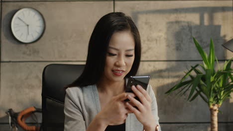 Close-Up-Of-The-Young-Pretty-Smiled-Businesswoman-Reading-Something-On-The-Smartphone-Screen-And-Texting-On-It-In-The-Office-And-Then-Looking-At-The-Side
