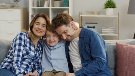 Portrait-Shot-Of-The-Young-Attractive-Parents-Sitting-With-Their-Little-Cute-Son-On-The-Sofa-In-The-Living-Room,-Hugging-And-Smiling-To-The-Camera-1