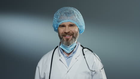 Porträtaufnahme-Des-Gutaussehenden-Arztes-Mit-Blauem-Hut,-Der-Die-Maske-Aus-Dem-Mund-Nimmt-Und-Fröhlich-In-Die-Kamera-Lächelt