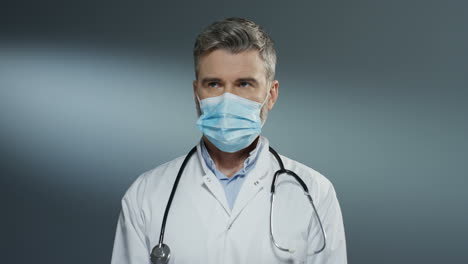 Gut-Aussehender-Chirurg,-Der-Eine-Blaue-Maske-Anzieht,-Wenn-Er-Sich-Für-Die-Operation-Auf-Grauem-Wandhintergrund-Vorbereitet
