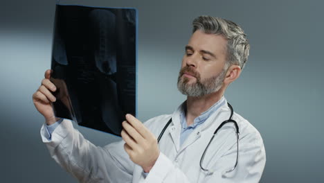 Grauhaariger,-Gut-Aussehender-Arzt,-Der-Eine-Röntgenkopie-Hält-Und-Sie-Sorgfältig-Auf-Dem-Grauen-Hintergrund-Studiert