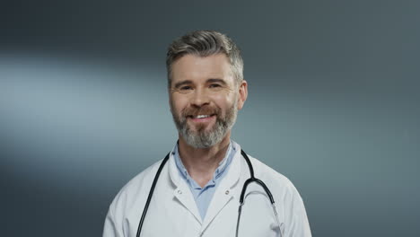 Porträtaufnahme-Des-Hübschen-Grauhaarigen-Männlichen-Arztes-Im-Weißen-Kittel-Und-Mit-Stethoskop-Am-Hals,-Der-Fröhlich-In-Die-Kamera-Lächelt